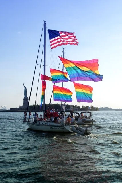 A sailing boat flying a United States flag and four rainbow flags from its masts and rigging, passing the Statue of Liberty in New York Harbor.