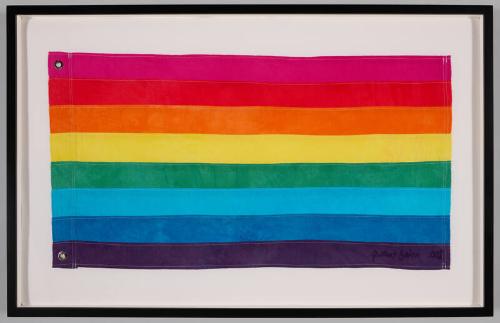 A framed eight-stripe hand-dyed cotton rainbow flag, hot pink at the top through violet at the bottom, signed by Gilbert Baker in the lower right corner.