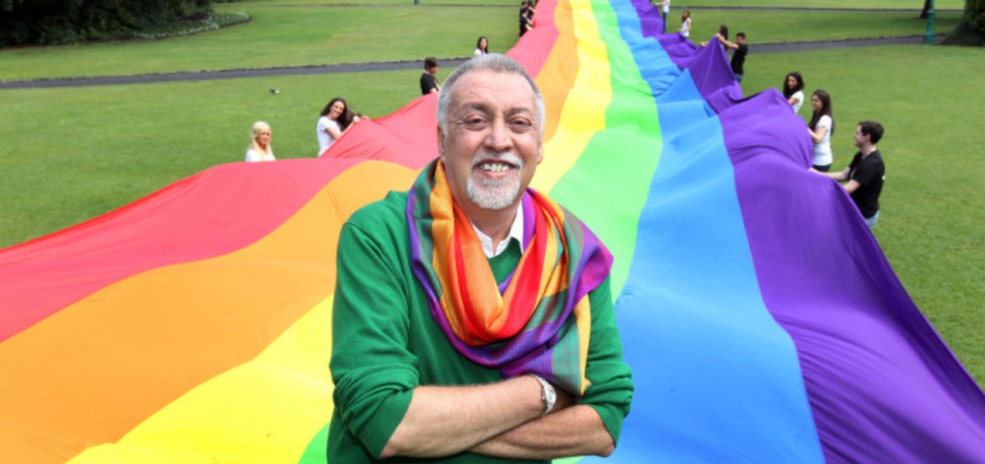 Gilbert Baker in a green sweater and rainbow scarf, arms crossed and smiling, standing before a giant six-stripe rainbow flag held by rows of volunteers in a park.