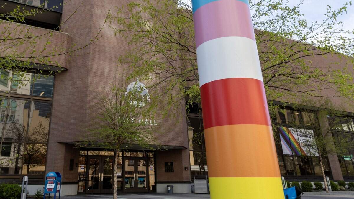 A City Hall flagpole wrapped in wide stripes of pink, white, blue, red, orange and yellow outside a brick building, with a rainbow window sign visible behind it.