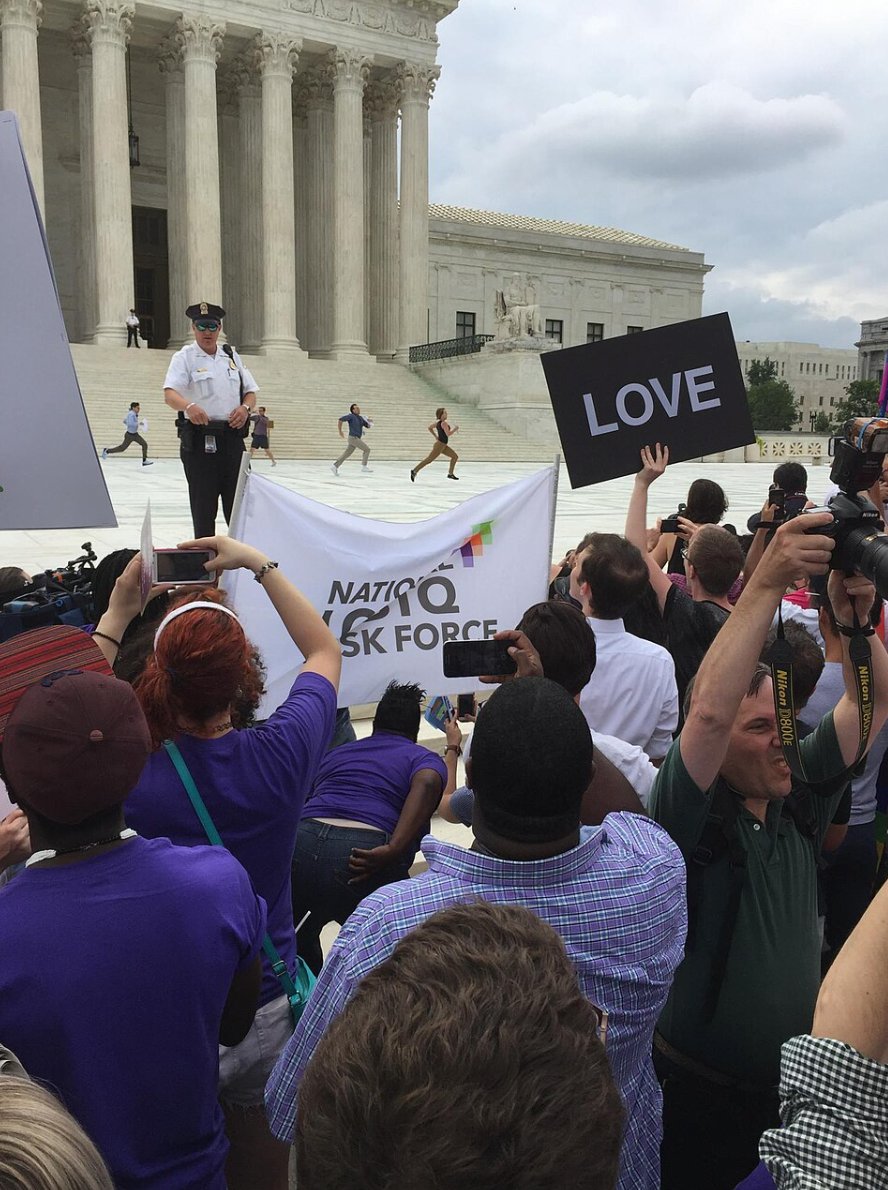 Interns sprinting across the plaza in front of the U.S. Supreme Court, one carrying a sheet of paper, watched by a police officer and a dense crowd holding a National LGBTQ Task Force banner and a placard reading LOVE.