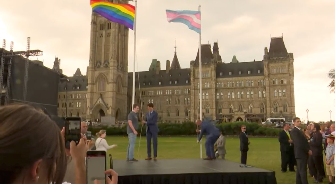On the lawn of Parliament Hill in Ottawa, a rainbow flag and a transgender pride flag fly from two masts while Prime Minister Justin Trudeau stands at the base of one, watched by a crowd holding up phones.