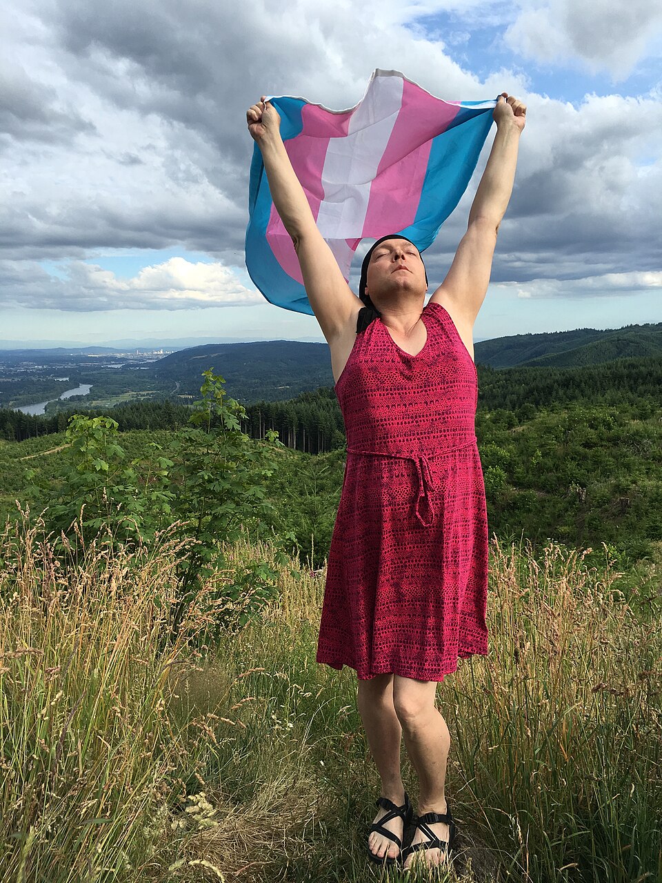 A person on a lawn holding a transgender pride flag spread open in both hands.