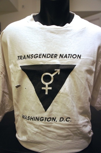 A white short-sleeved T-shirt displayed on a black mannequin form. Arched across the chest in black block capitals: TRANSGENDER NATION. Below it a solid black downward-pointing triangle, and reversed out of the triangle in white a single combined transgender symbol, a circle with a cross descending from it and an arrow rising to the upper right. Beneath the triangle, in black capitals: WASHINGTON, D.C. A small tear is visible at the left edge of the shirt.