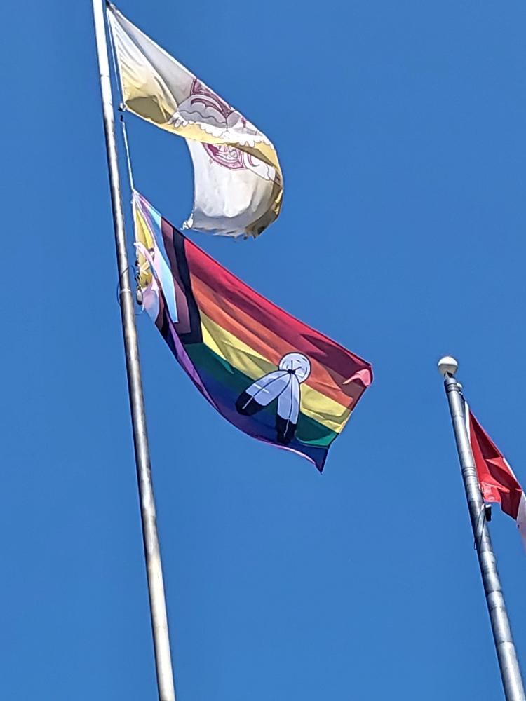 A Pride flag flying from a pole against a blue sky: six rainbow stripes with a hoist chevron and yellow field, carrying two white feathers with black tips bound beneath a white circle.