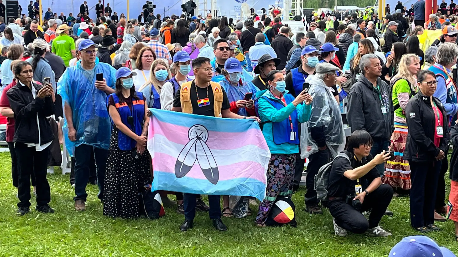 A cloth flag with the five stripes of the Transgender Pride Flag, charged with two feathers hanging beneath a circle, held open at chest height in a crowd standing on grass, many in rain ponchos.