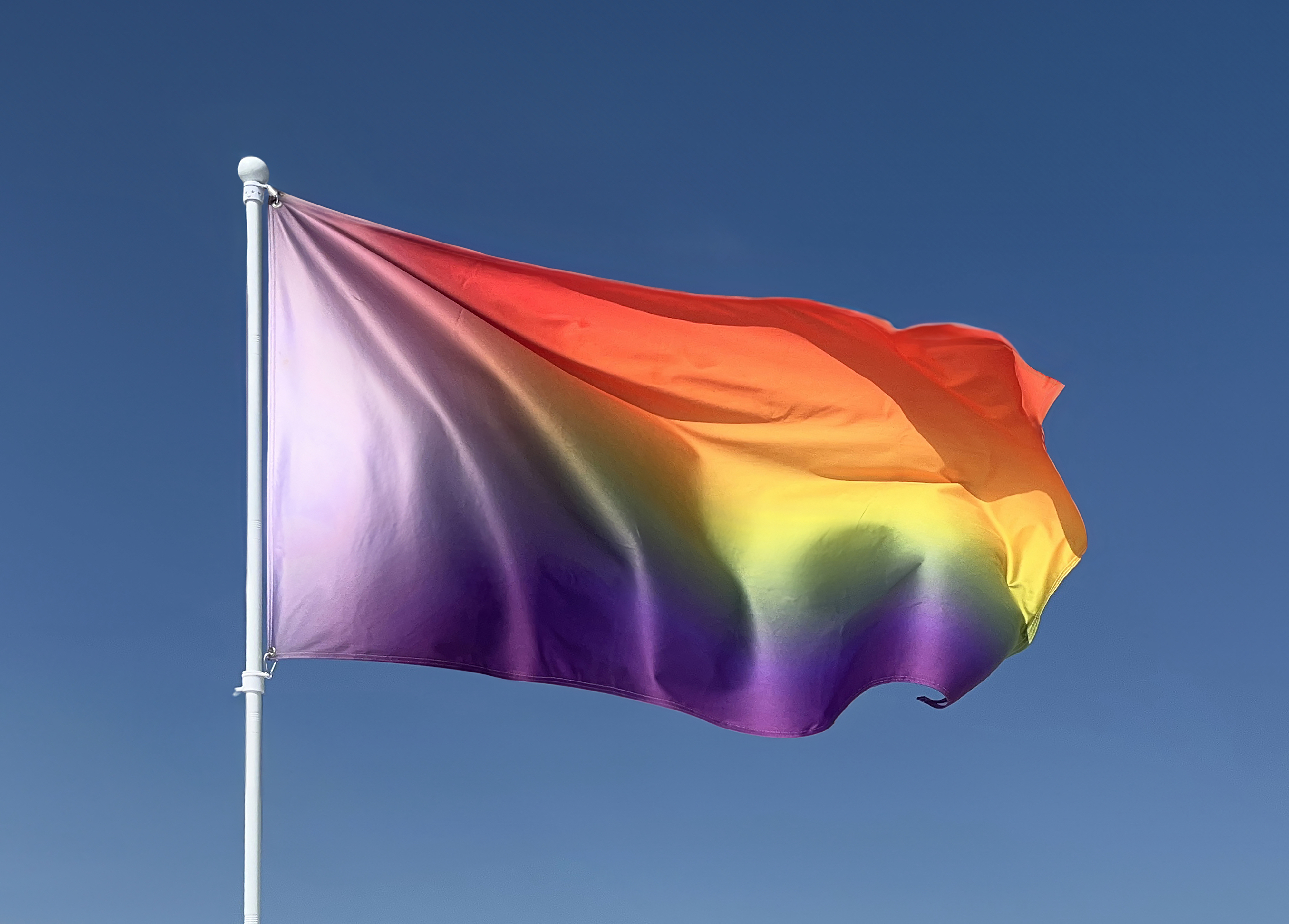 Utopian Pride Flag #6 flying from a white flagpole against a clear blue sky, its blurred rainbow running red at the top through yellow to purple, with the pale chevron region at the hoist.