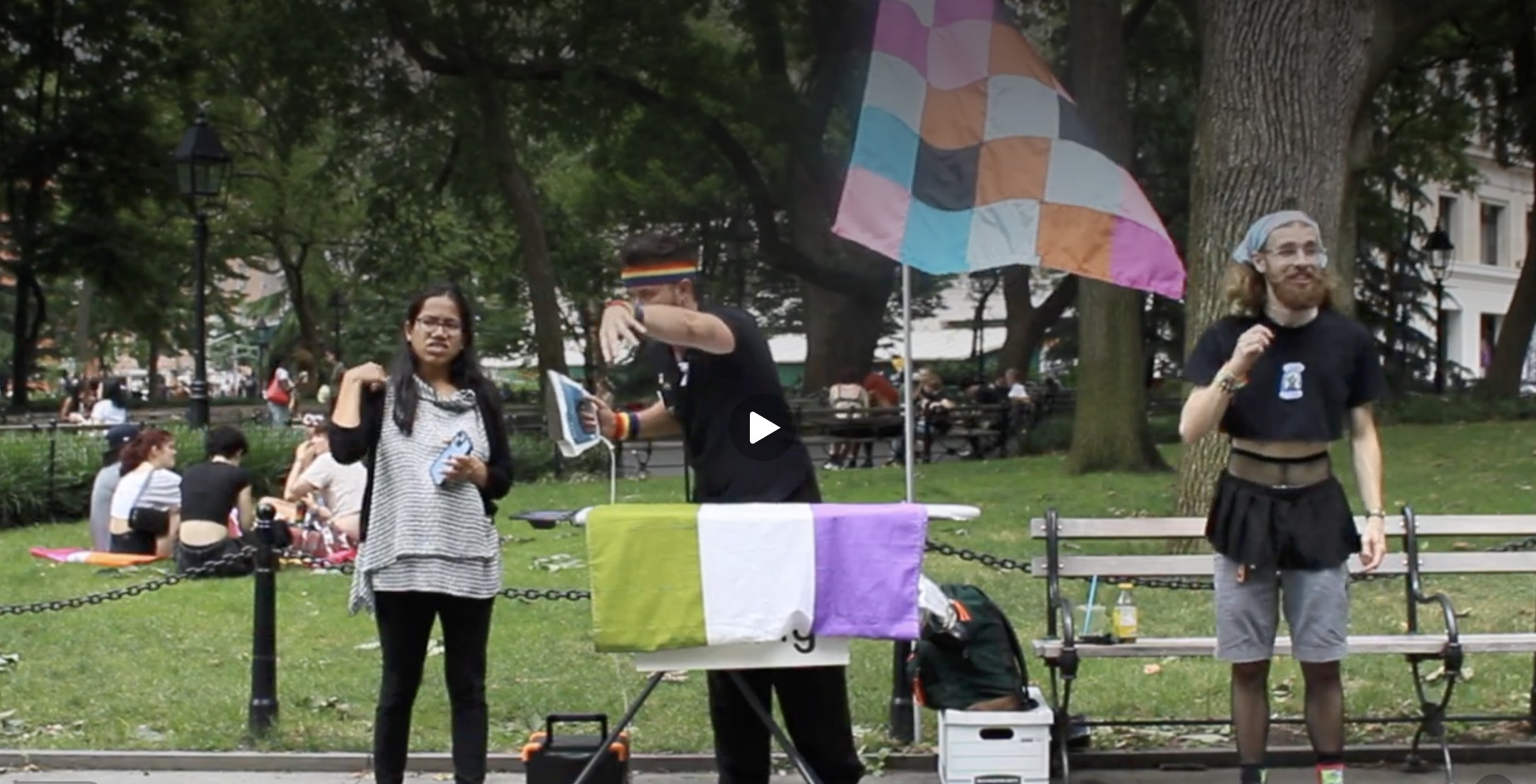 Three people beside an ironing board in a park. A flag with green, white and lavender stripes is spread across the board; a Progress Pride flag flies behind them.