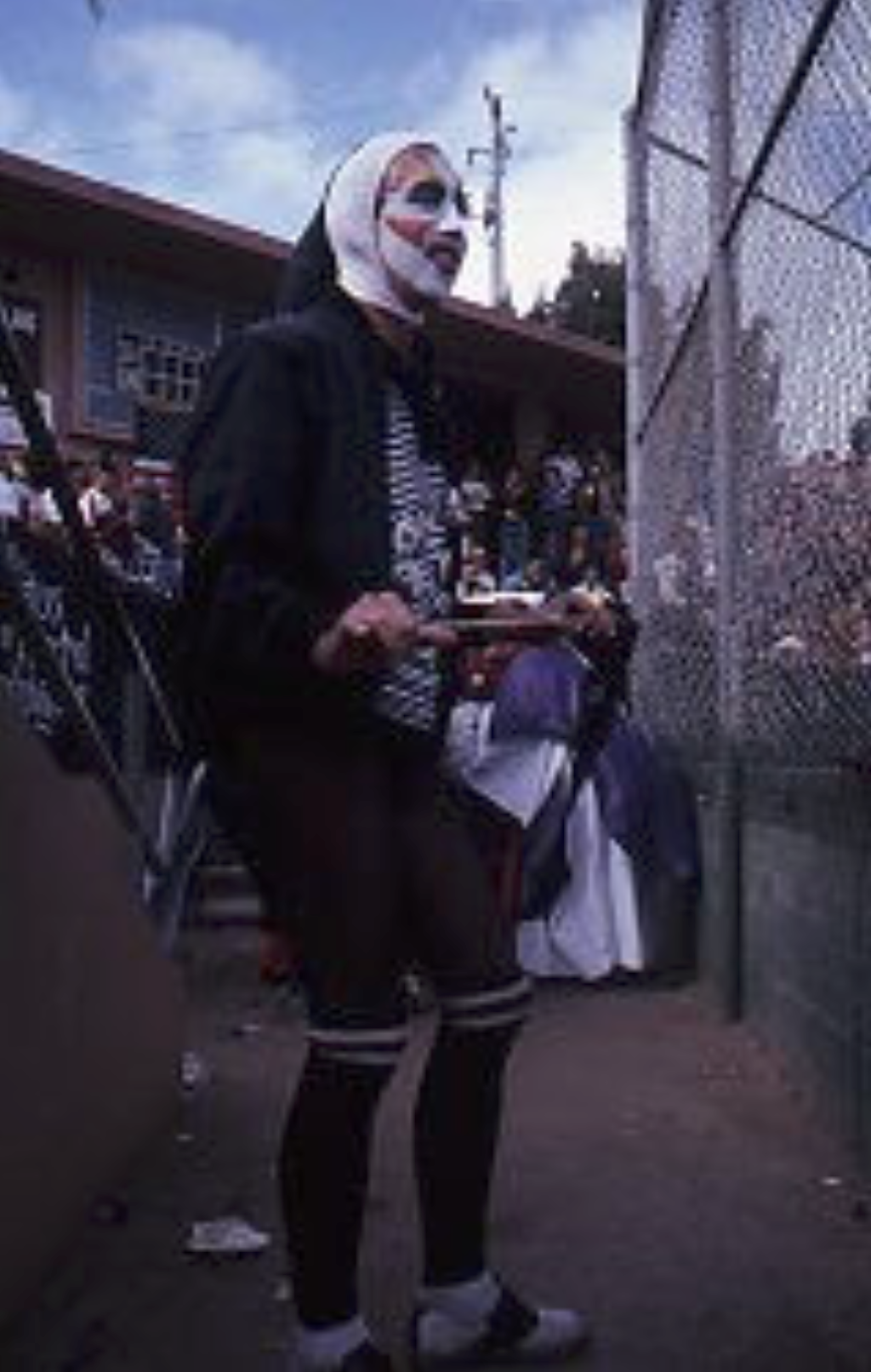 Gilbert Baker in a nun's habit and whiteface makeup with black eye and mouth markings, standing by a chain-link backstop at a softball game.