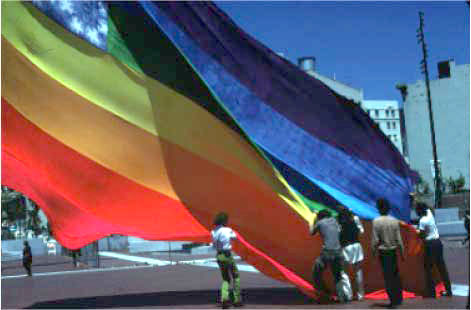 Several people standing beneath one of the original 1978 rainbow flags, holding its lower edge as it billows above a paved plaza.